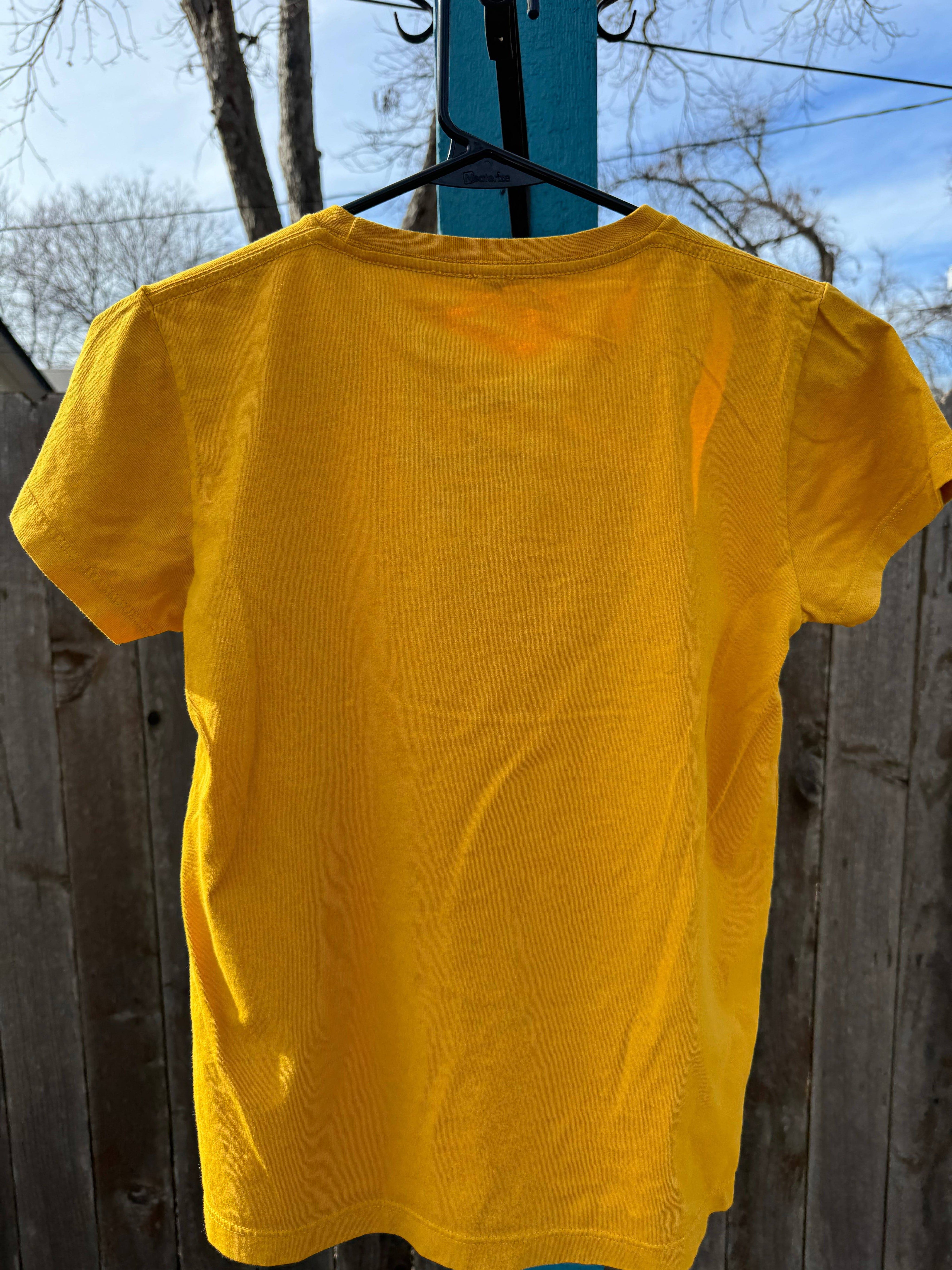 A yellow Kids OG Bison Tee by Farm Fresh Clothing, made of organic cotton in the USA, hangs on a black hanger outdoors with its back facing the camera, set against a wooden fence and leafless trees under a blue sky.