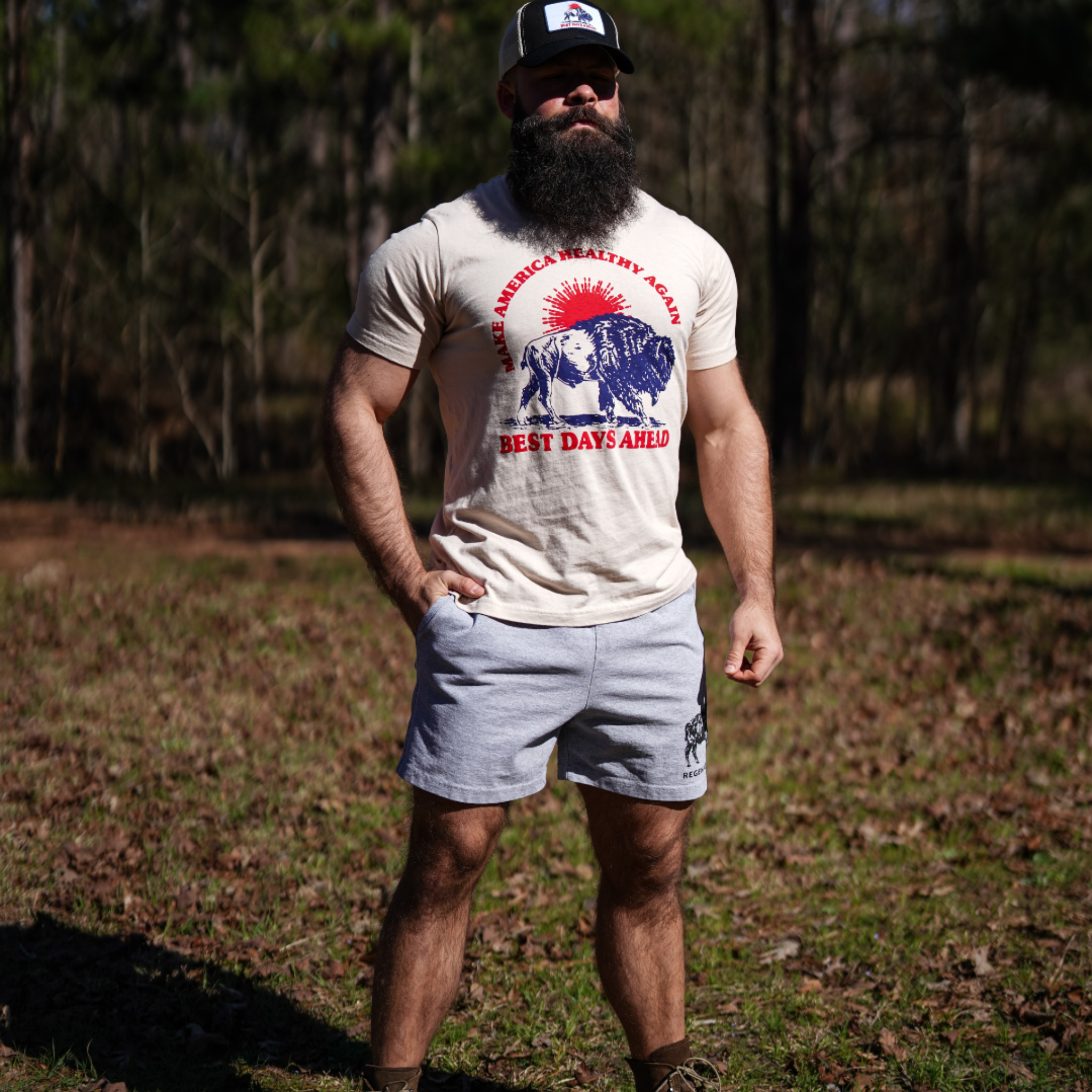 A bearded man in a cap and Farm Fresh Clothings Best Days Ahead MAHA Tee in sand stands outdoors on a sunny day, with trees behind him, embodying the spirit of regenerative agriculture.