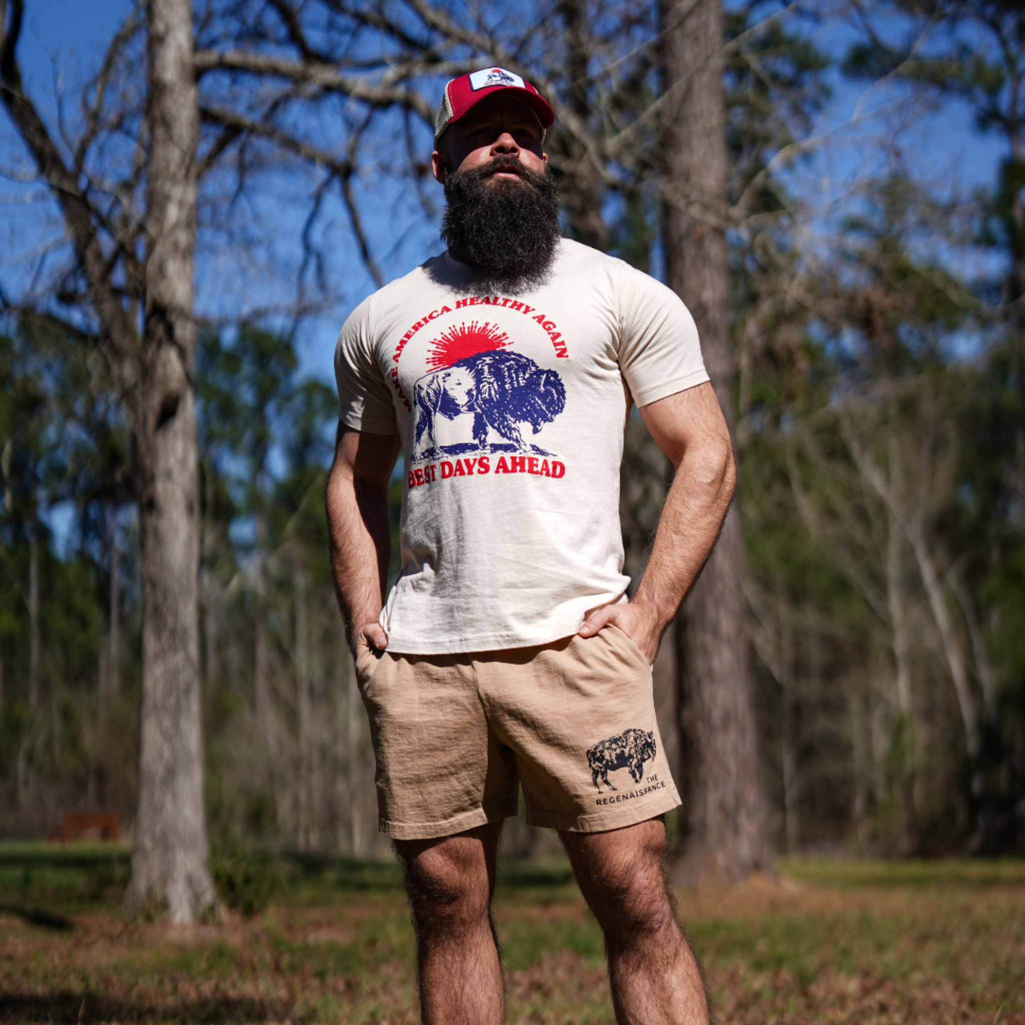 A bearded man in a cap and eggshell Best Days Ahead MAHA Tee by Farm Fresh Clothing stands confidently among tall sunlit trees, embodying the brand’s commitment to regenerative agriculture and a deep appreciation for the land.