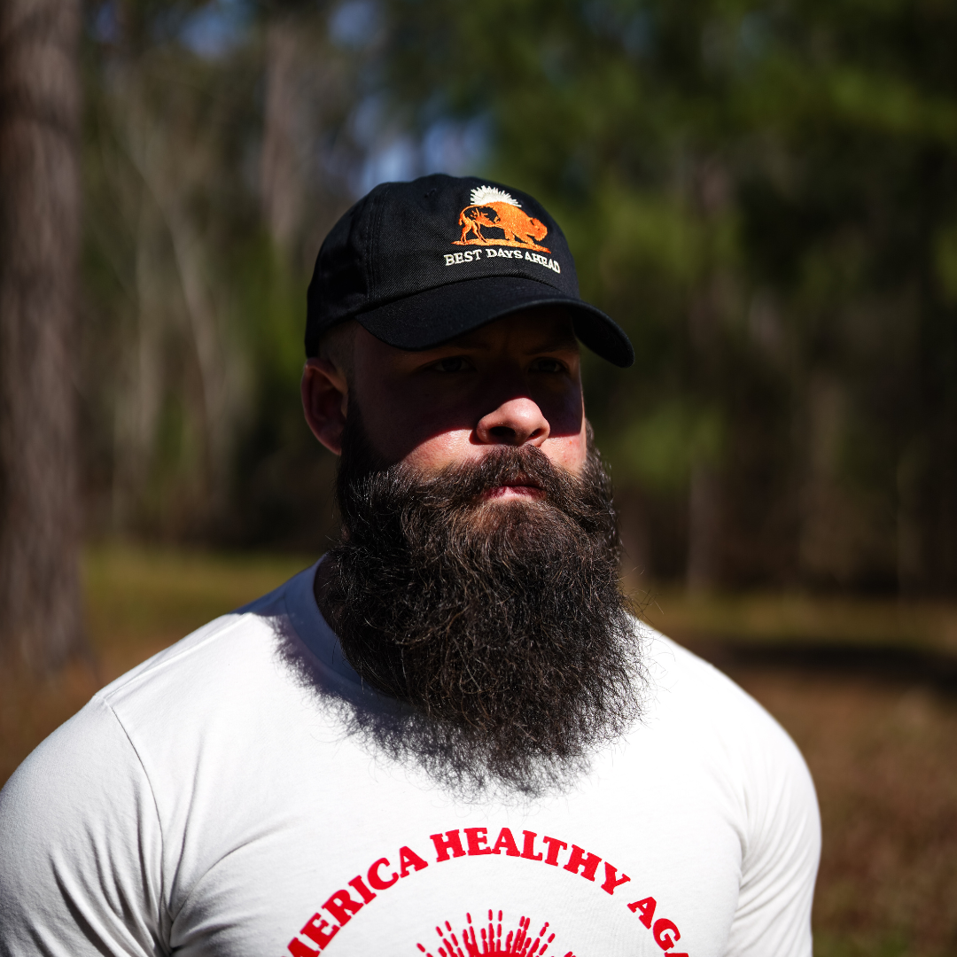 A bearded man stands outdoors in a sunlit, wooded area wearing a black Best Days Ahead MAHA Cap by Farm Fresh Clothing and a white Make America Healthy Again T-shirt, embracing regenerative health.