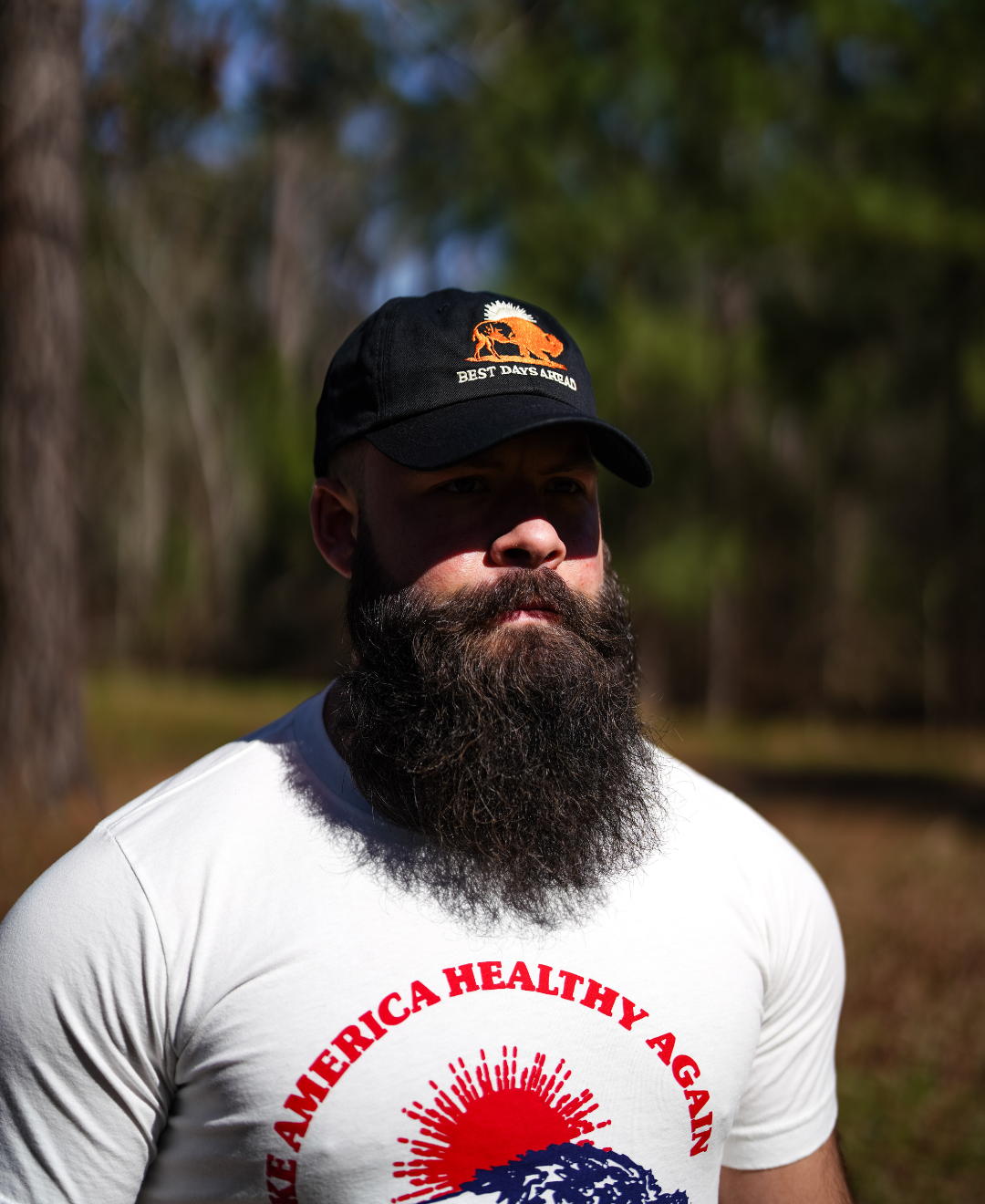 A bearded man stands outdoors in a sunlit, wooded area wearing a black Best Days Ahead MAHA Cap by Farm Fresh Clothing and a white Make America Healthy Again T-shirt, embracing regenerative health.