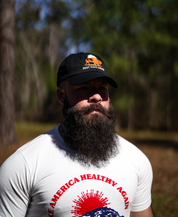 A bearded man stands outdoors in a sunlit, wooded area wearing a black Best Days Ahead MAHA Cap by Farm Fresh Clothing and a white Make America Healthy Again T-shirt, embracing regenerative health.