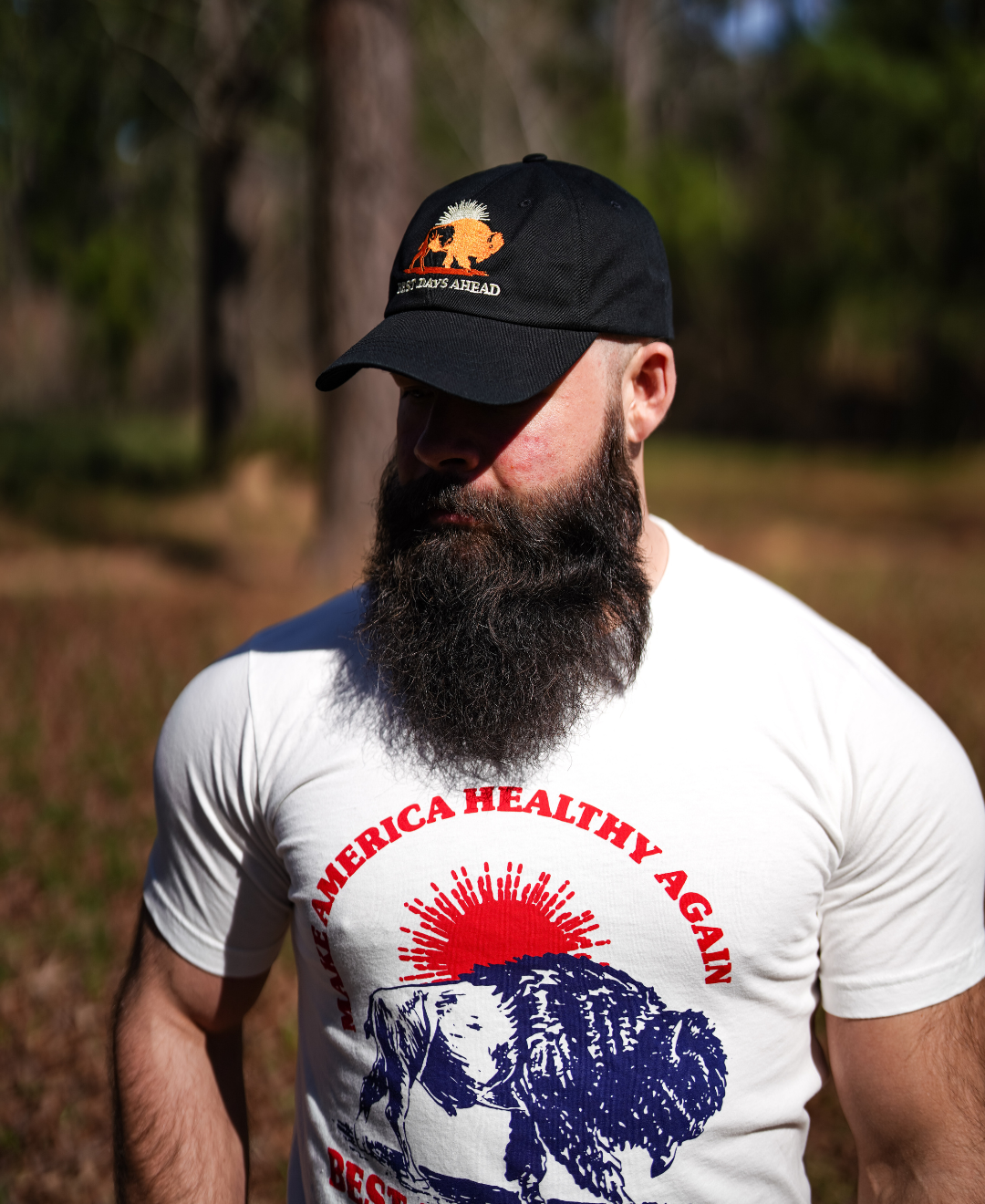 A bearded man in a Farm Fresh Clothing Best Days Ahead MAHA Cap and a white t-shirt with a bison graphic and Make America Healthy Again stands outdoors in a sunlit, wooded area, capturing the spirit of regenerative health.