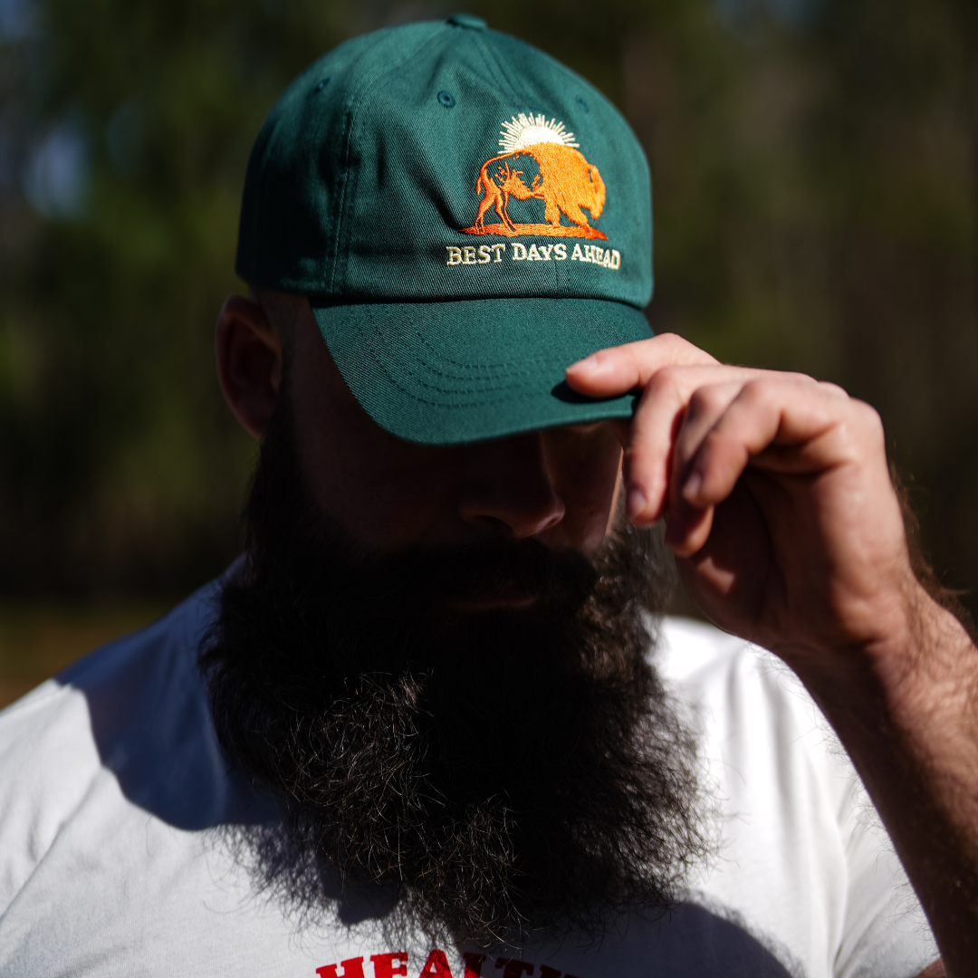 A bearded man outdoors adjusts the brim of his spruce Best Days Ahead MAHA Cap from Farm Fresh Clothing, embracing regenerative health in a white tee with red lettering.