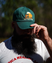 A bearded man outdoors adjusts the brim of his spruce Best Days Ahead MAHA Cap from Farm Fresh Clothing, embracing regenerative health in a white tee with red lettering.