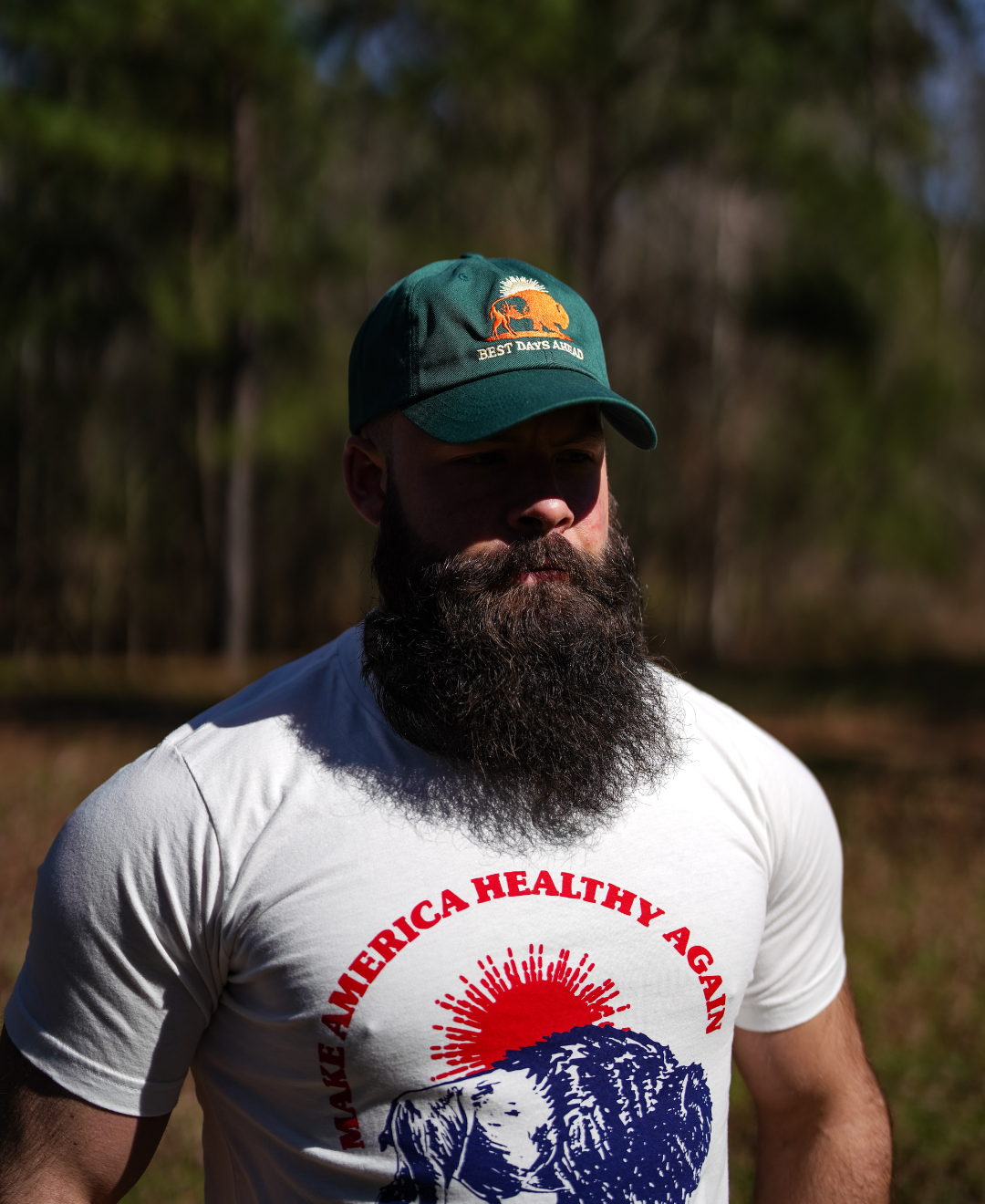A bearded man wears a white “Make America Healthy Again” shirt and a Farm Fresh Clothing Best Days Ahead MAHA Cap as he stands outdoors in a sunlit, wooded area, embodying regenerative health.