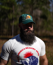A bearded man wears a white “Make America Healthy Again” shirt and a Farm Fresh Clothing Best Days Ahead MAHA Cap as he stands outdoors in a sunlit, wooded area, embodying regenerative health.