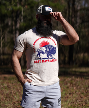 A bearded man in a cap, sunglasses, and a “Make America Healthy Again—Best Days Ahead” tee stands on grass with trees behind him, holding the brim of his Farm Fresh Clothing Best Days Ahead MAHA Trucker Hat in Charcoal/Oyster.
