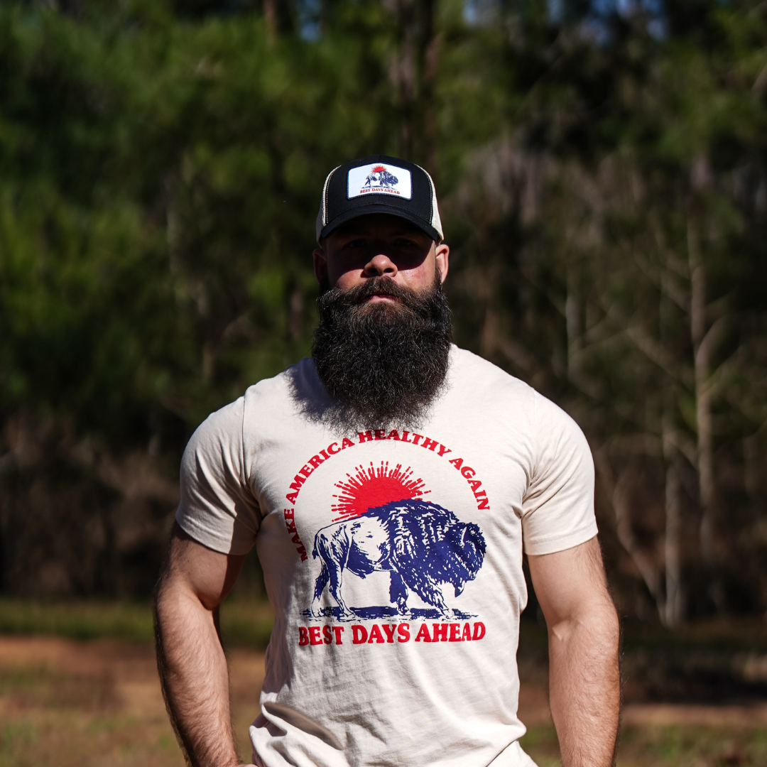 A bearded man stands outdoors among pine trees, wearing the Farm Fresh Clothing Best Days Ahead MAHA Trucker Hat and a T-shirt with a lion, child, and the words MAKE AMERICA HEALTHY AGAIN BEST DAYS AHEAD.