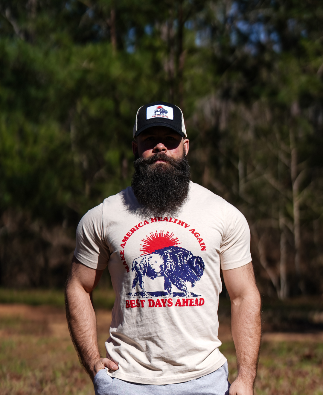 A bearded man stands outdoors among pine trees, wearing the Farm Fresh Clothing Best Days Ahead MAHA Trucker Hat and a T-shirt with a lion, child, and the words MAKE AMERICA HEALTHY AGAIN BEST DAYS AHEAD.