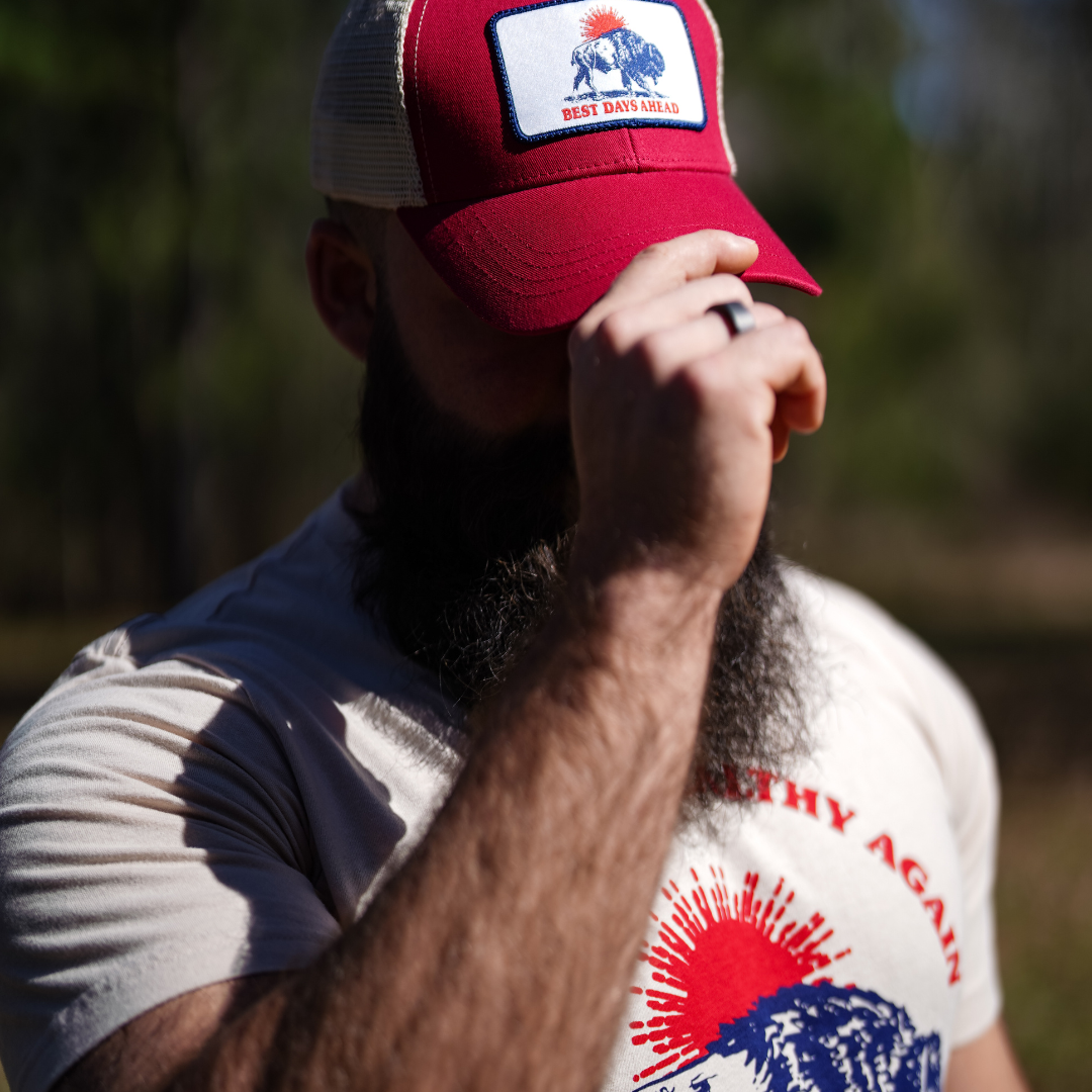 oyster Best Days Ahead MAHA Trucker Hat by Farm Fresh Clothing stands outdoors, partially shielding their face with a hand. Sunlit trees blur in the background, evoking the promise of nature-filled days ahead.