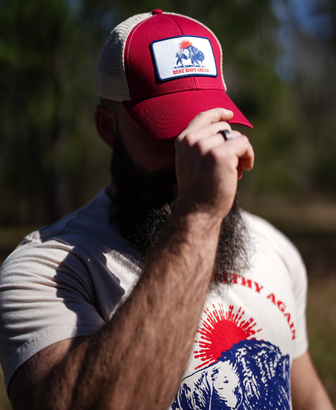 A bearded person in a red/oyster Best Days Ahead MAHA Trucker Hat by Farm Fresh Clothing stands outdoors, partially shielding their face with a hand. Sunlit trees blur in the background, evoking the promise of nature-filled days ahead.