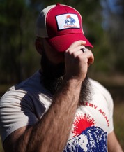 A bearded person in a red/oyster Best Days Ahead MAHA Trucker Hat by Farm Fresh Clothing stands outdoors, partially shielding their face with a hand. Sunlit trees blur in the background, evoking the promise of nature-filled days ahead.