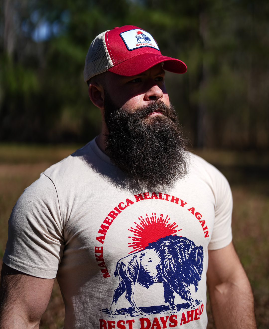 A bearded man wears the Farm Fresh Clothing Best Days Ahead MAHA Trucker Hat and a Make America Healthy Again, Best Days Ahead bison T-shirt, standing outdoors in sunlight with trees—embracing regenerative agriculture.