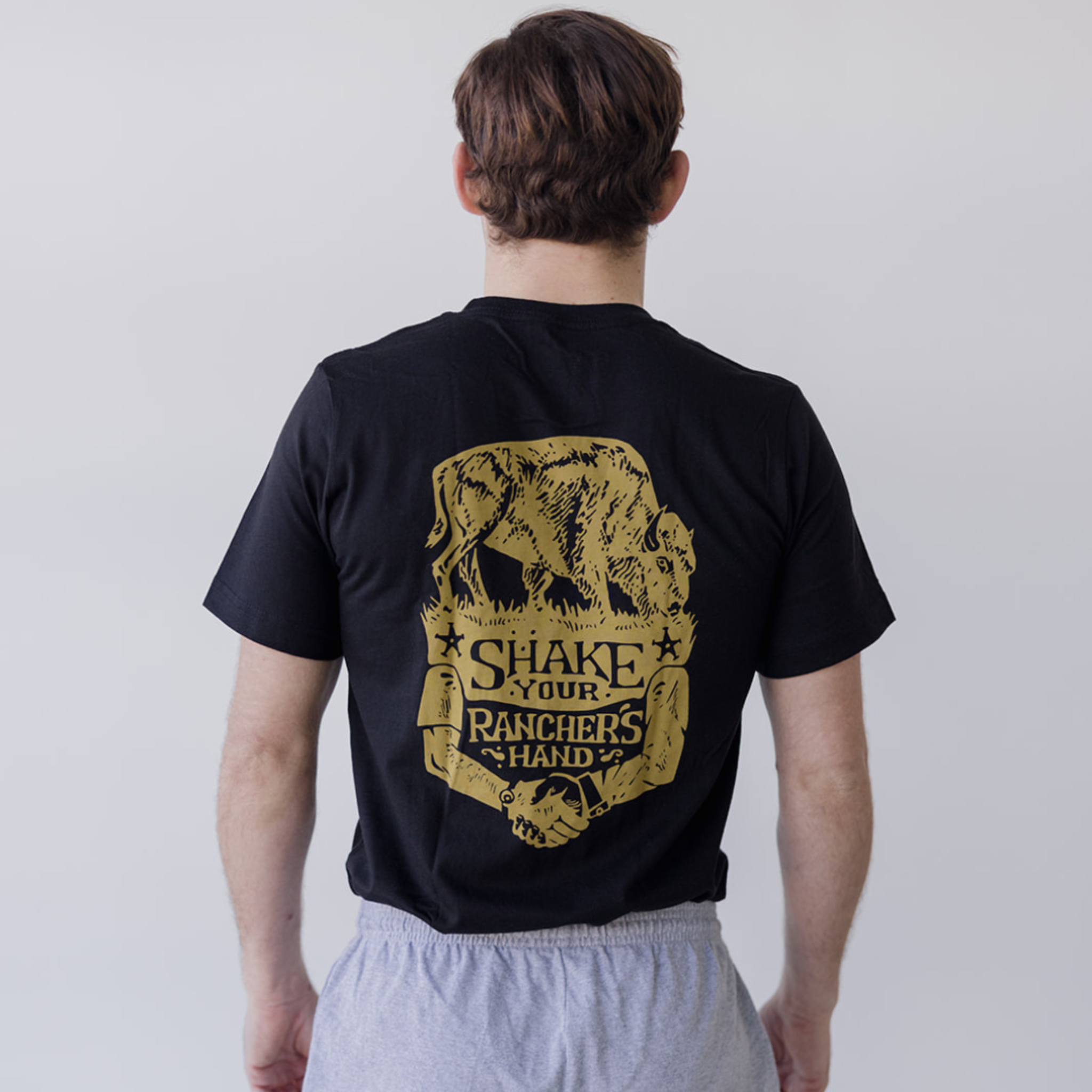 A person in Farm Fresh Clothing’s Bison Ranchers Handshake Tee—organic cotton with a gold bison graphic and Shake Your Ranchers Hand on the back—stands before a plain light background, promoting transparent food systems.