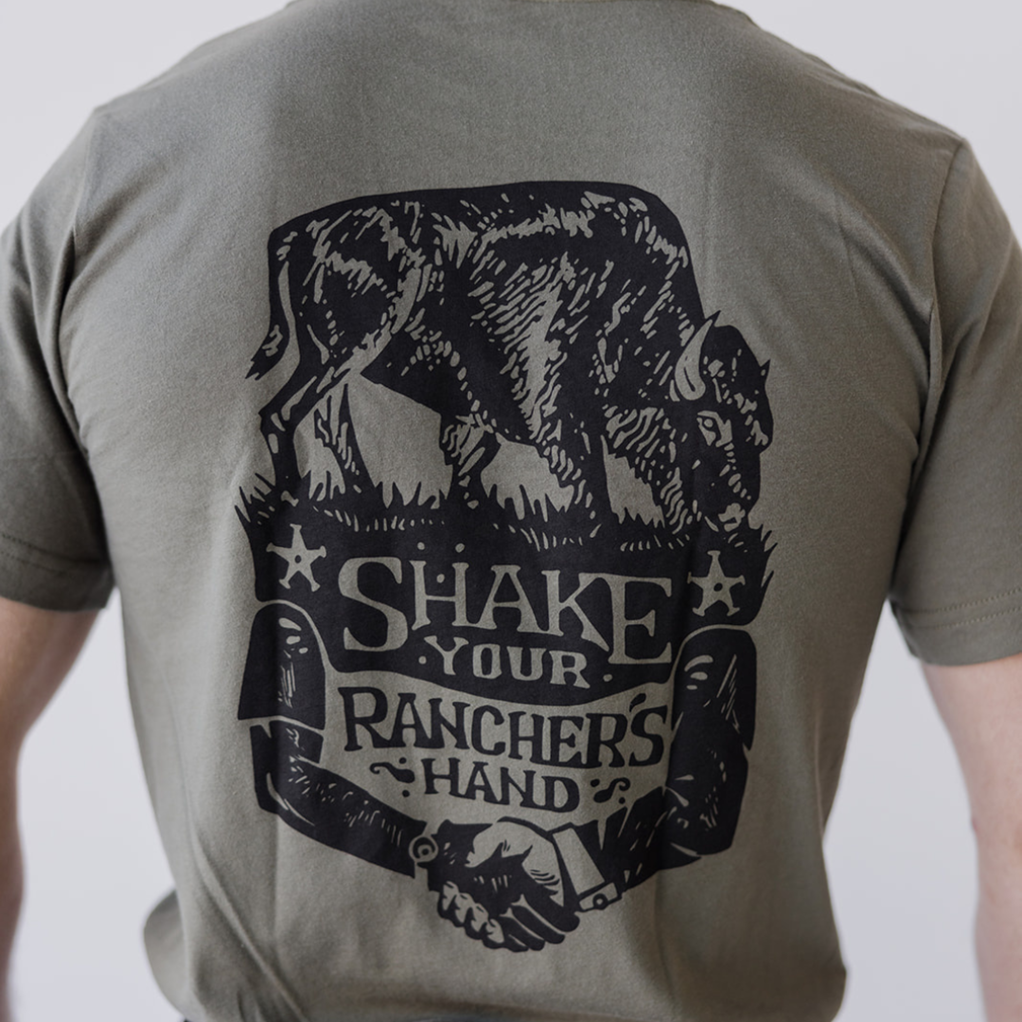 A person wears the Farm Fresh Clothing Bison Ranchers Handshake Tee, made from organic cotton, with a black cow-on-grass graphic above Shake Your Ranchers Hand and two hands shaking—ideal for transparent food system fans.