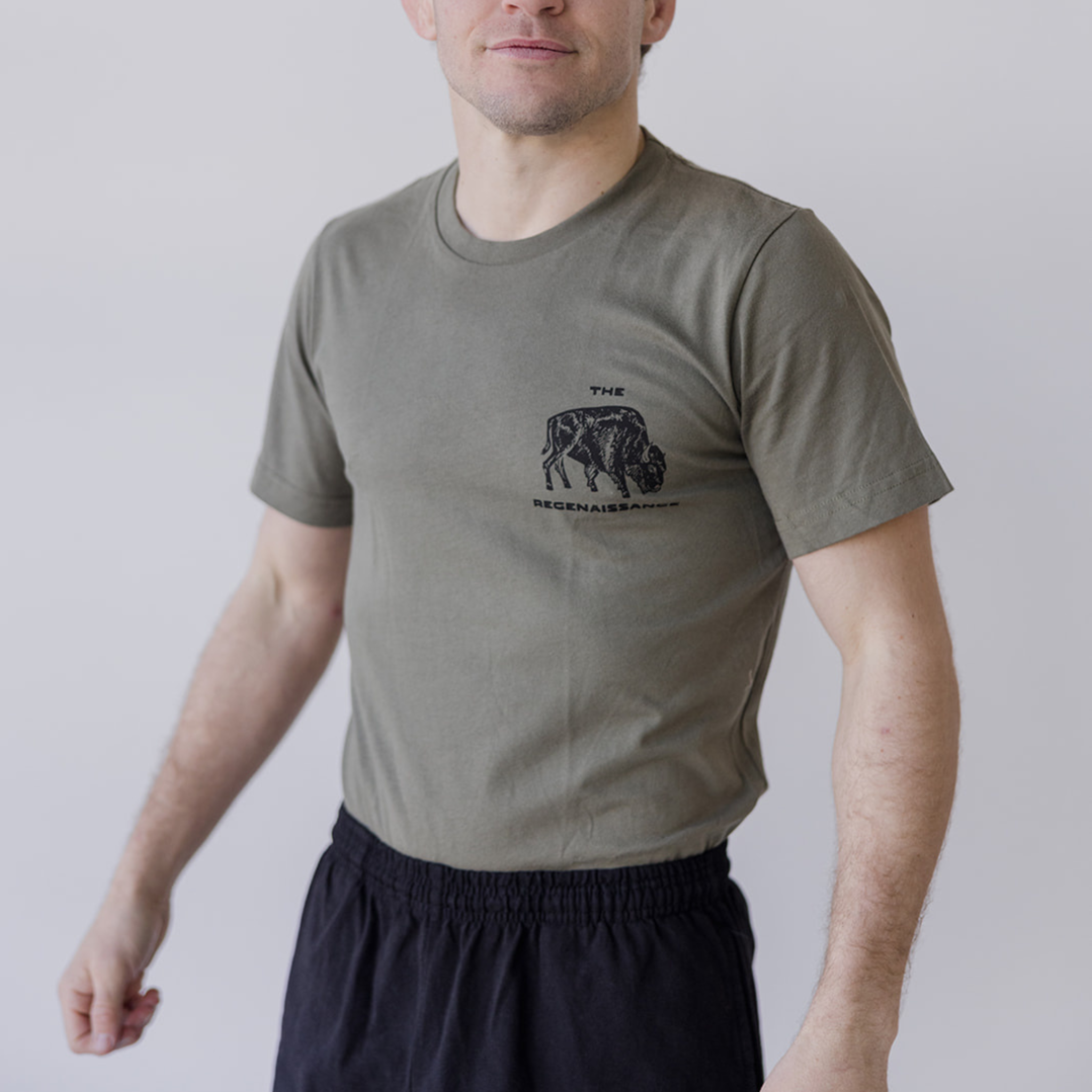 A person wears the Farm Fresh Clothing Bison Ranchers Handshake Tee in rosemary with a buffalo graphic and THE REGENASSANCE on the front left chest, paired with black elastic-waist shorts, standing against a plain light background.