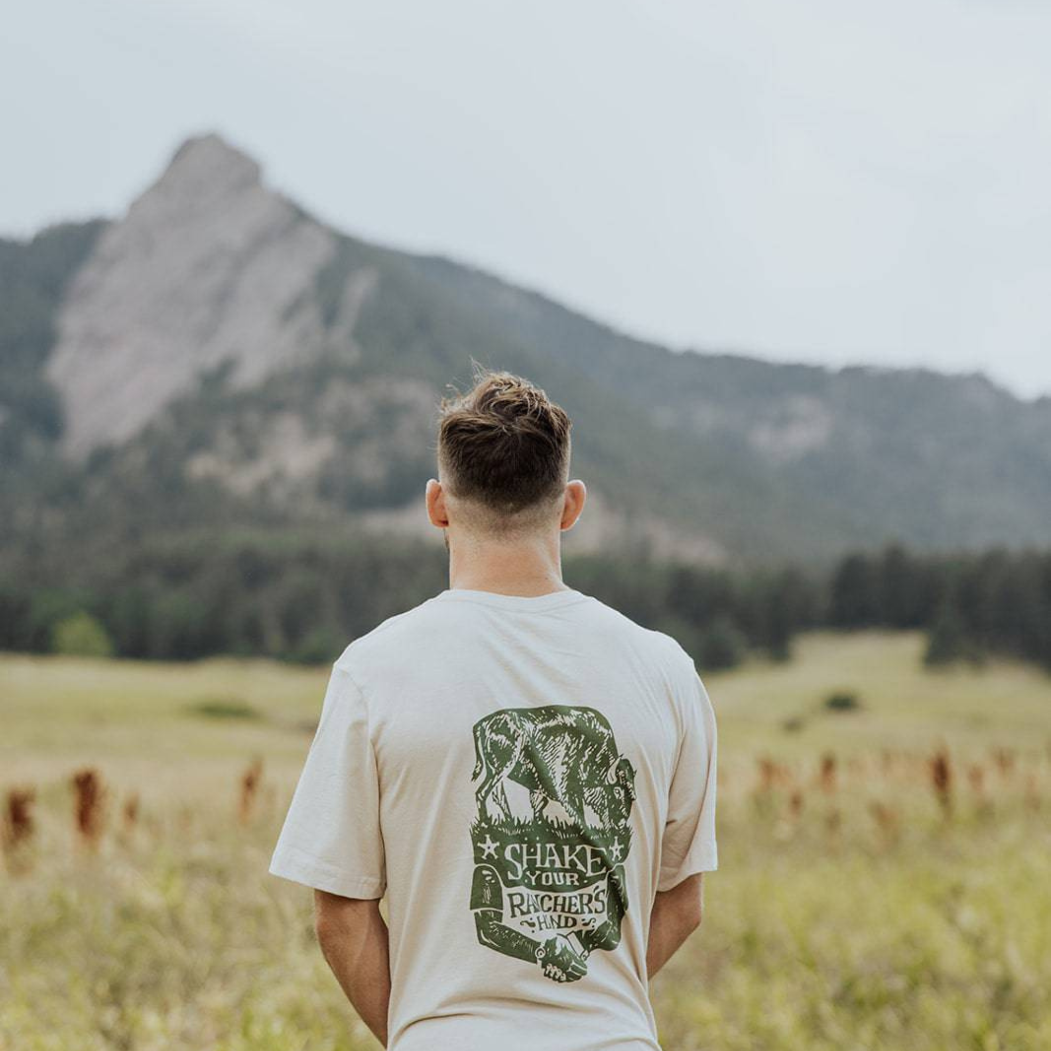 In a grassy field under cloudy skies, a person faces a distant mountain wearing the Farm Fresh Clothing Bison Ranchers Handshake Tee in Sand, made with organic cotton and featuring “Shake Your Riches Wind” on the back, evoking transparent food systems.