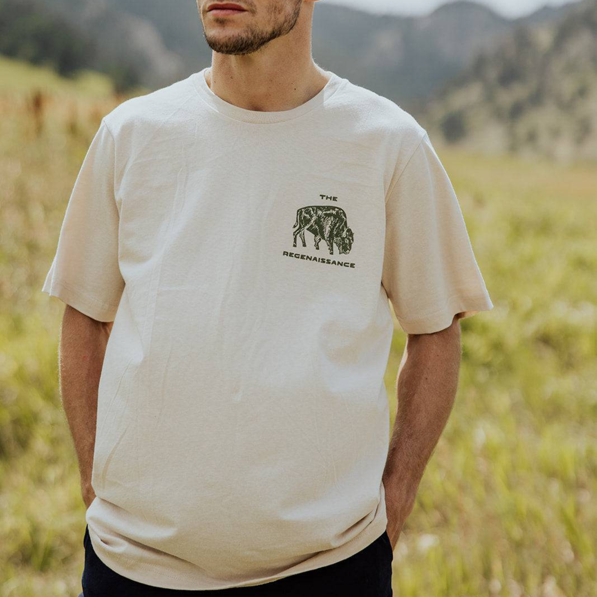 A man stands in a grassy, mountainous landscape wearing the Farm Fresh Clothing Bison Ranchers Handshake Tee featuring a small rhinoceros graphic and the text The Reconnaissance.