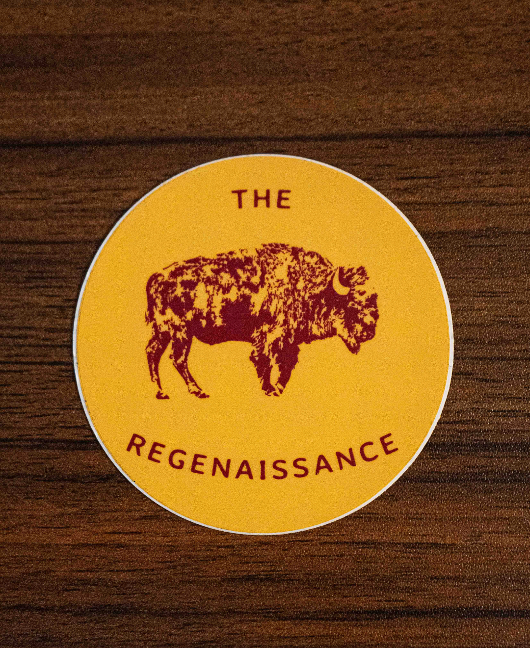 The Regenaissance Sticker by Sticker Mule is a round yellow vinyl sticker with a maroon Great American Bison and THE REGENAISSANCE in maroon, shown on a wooden surface.