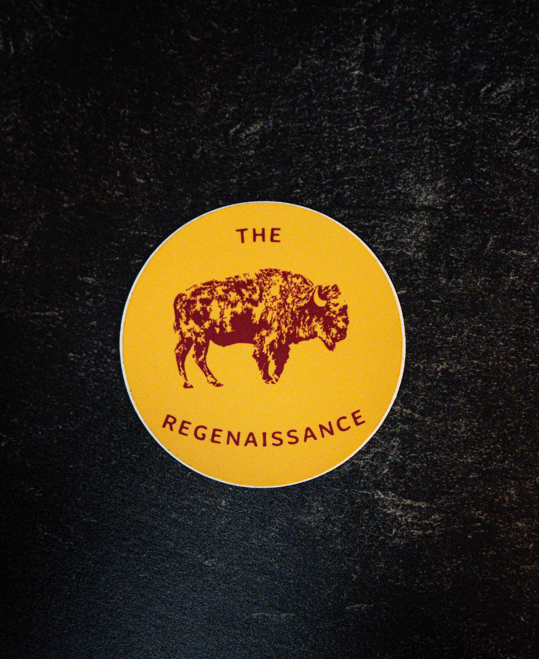 The Regenaissance Sticker by Sticker Mule is a circular yellow vinyl sticker with a maroon Great American Bison illustration and THE REGENAISSANCE in maroon, shown on a dark textured background.