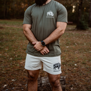 A bearded man in an olive green tee and Los Angeles Apparel Men's Bison Shorts with a black animal graphic stands on grass, hands clasped and wearing a smartwatch. Trees and fallen leaves evoke his regenerative lifestyle.