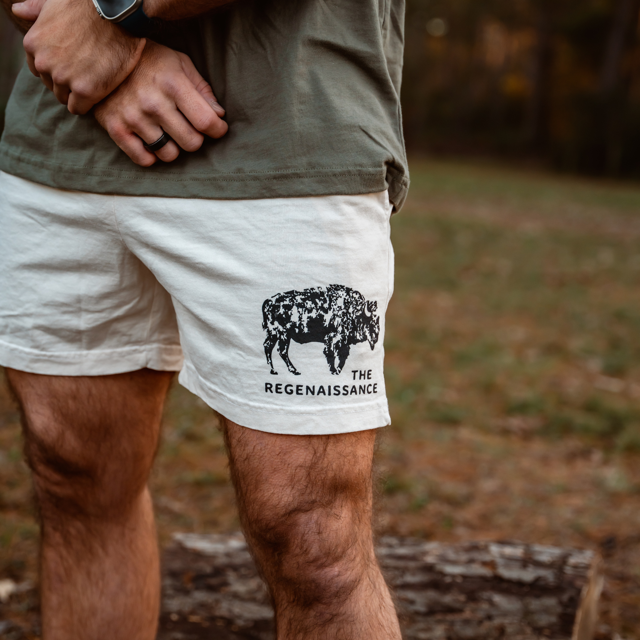 Outdoors, a person wears Los Angeles Apparel Men's Bison Shorts—white with a black bison illustration and THE REGENAISSANCE logo—reflecting regenerative living. The grassy background is gently blurred.