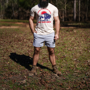 A bearded person stands outdoors on grass and leaves, wearing a beige "Best Days Ahead" cow and red sun tee, Los Angeles Apparel Men's Bison Shorts in gray, and brown boots—reflecting the spirit of regenerative agriculture. Trees are behind.