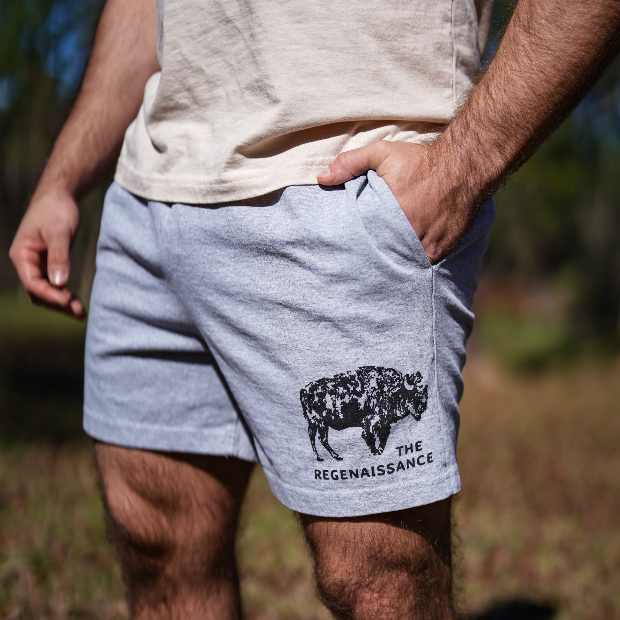 A person stands outdoors in Los Angeles Apparel Men's Bison Shorts, with a black bison graphic and "The Regenaissance" on the leg. One hand is in a pocket as trees and grass blur gently in the background.