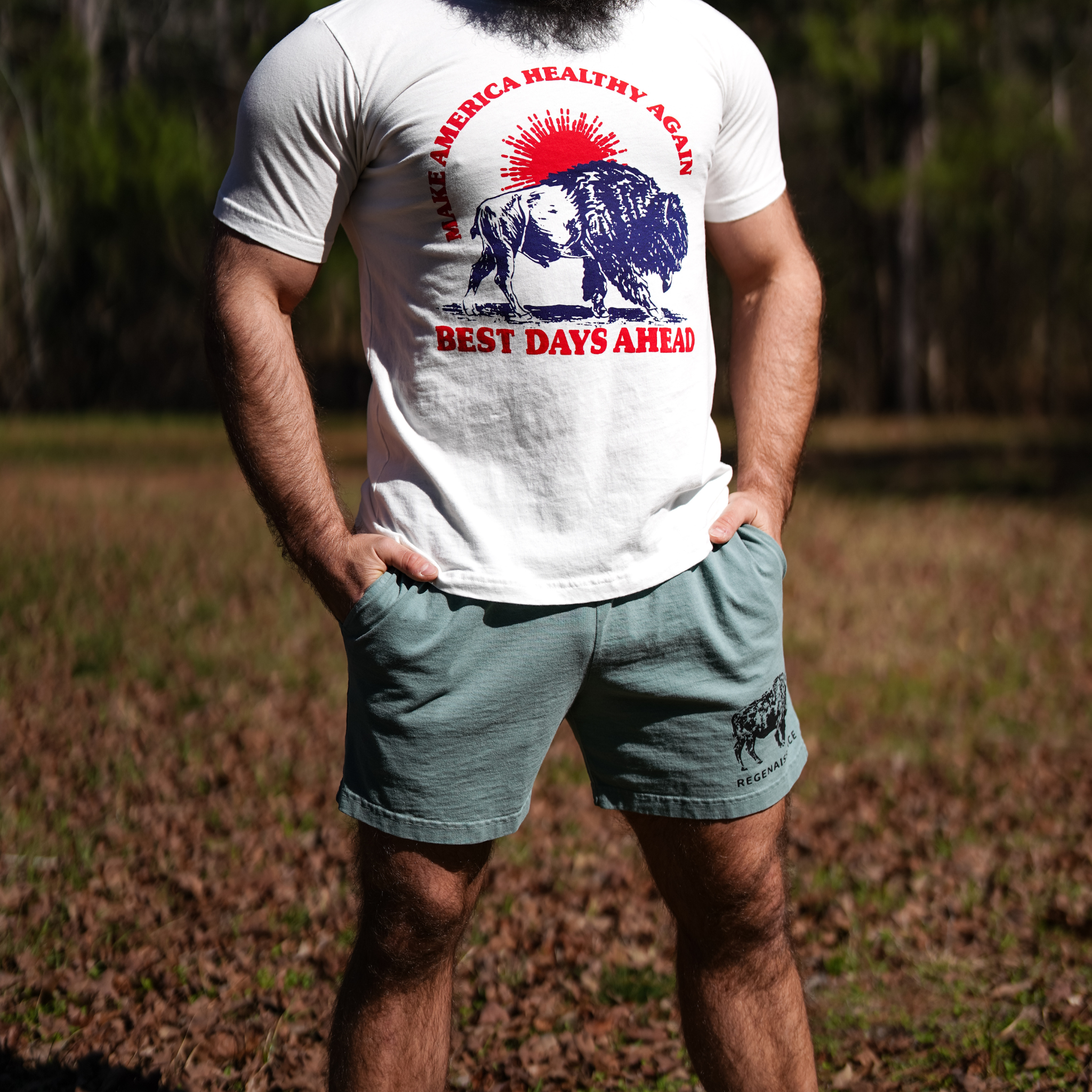 A bearded man stands on grass wearing Los Angeles Apparel's Men's Bison Shorts and a white T-shirt with a buffalo graphic and "Make America Healthy Again – Best Days Ahead"; trees are visible in the background, evoking regenerative living.