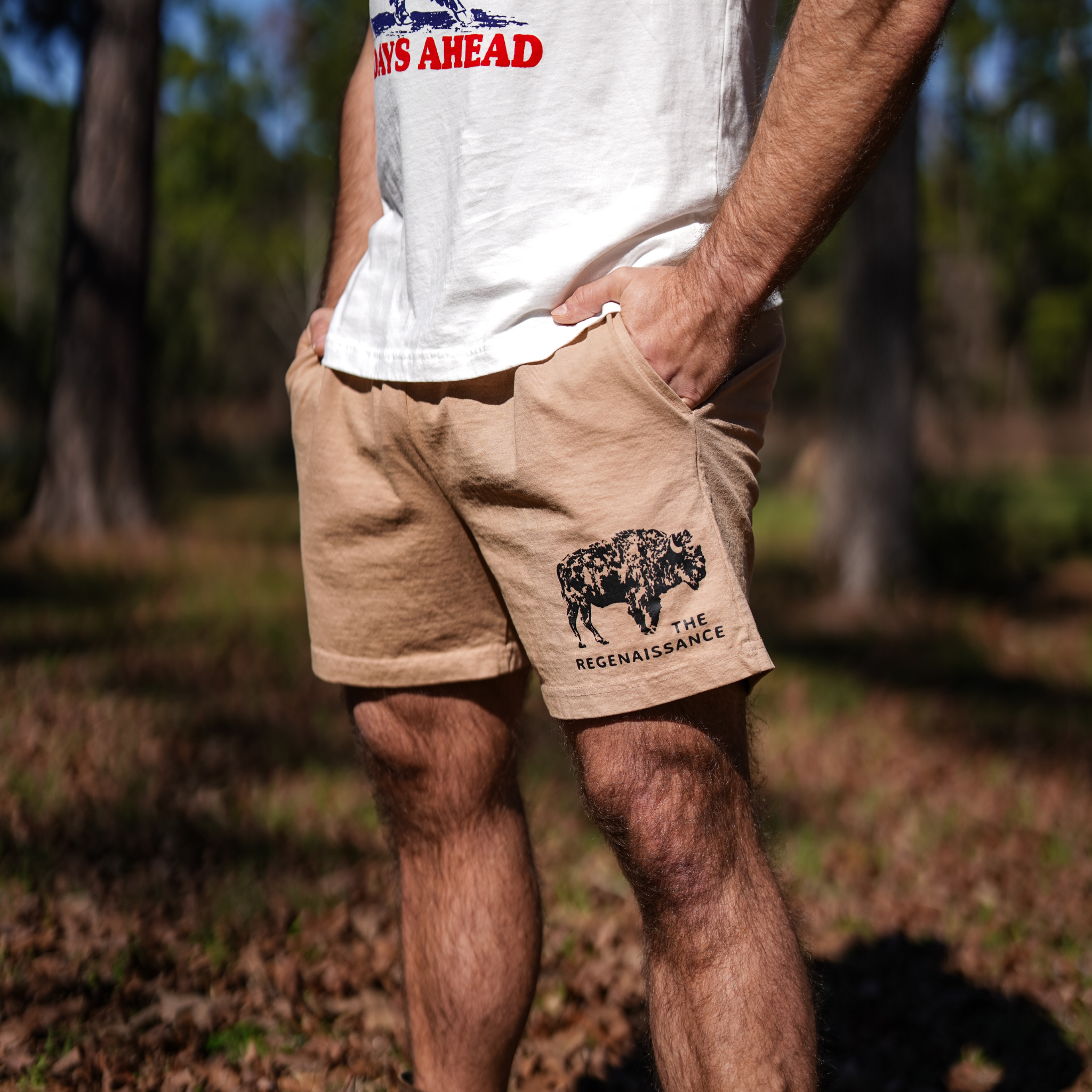 Wearing Los Angeles Apparel Men's Bison Shorts, featuring a black bison graphic and "THE REGENAISSANCE" on the left leg, this person stands with hands in pockets in a white shirt amid trees and grass, embodying regenerative living.