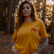 A woman with wavy hair and tattoos poses outdoors in a sunlit forest wearing the Farm Fresh Clothing Bison Tee, a mustard organic cotton T-shirt featuring red The Renaissance text and an illustration of two lions.