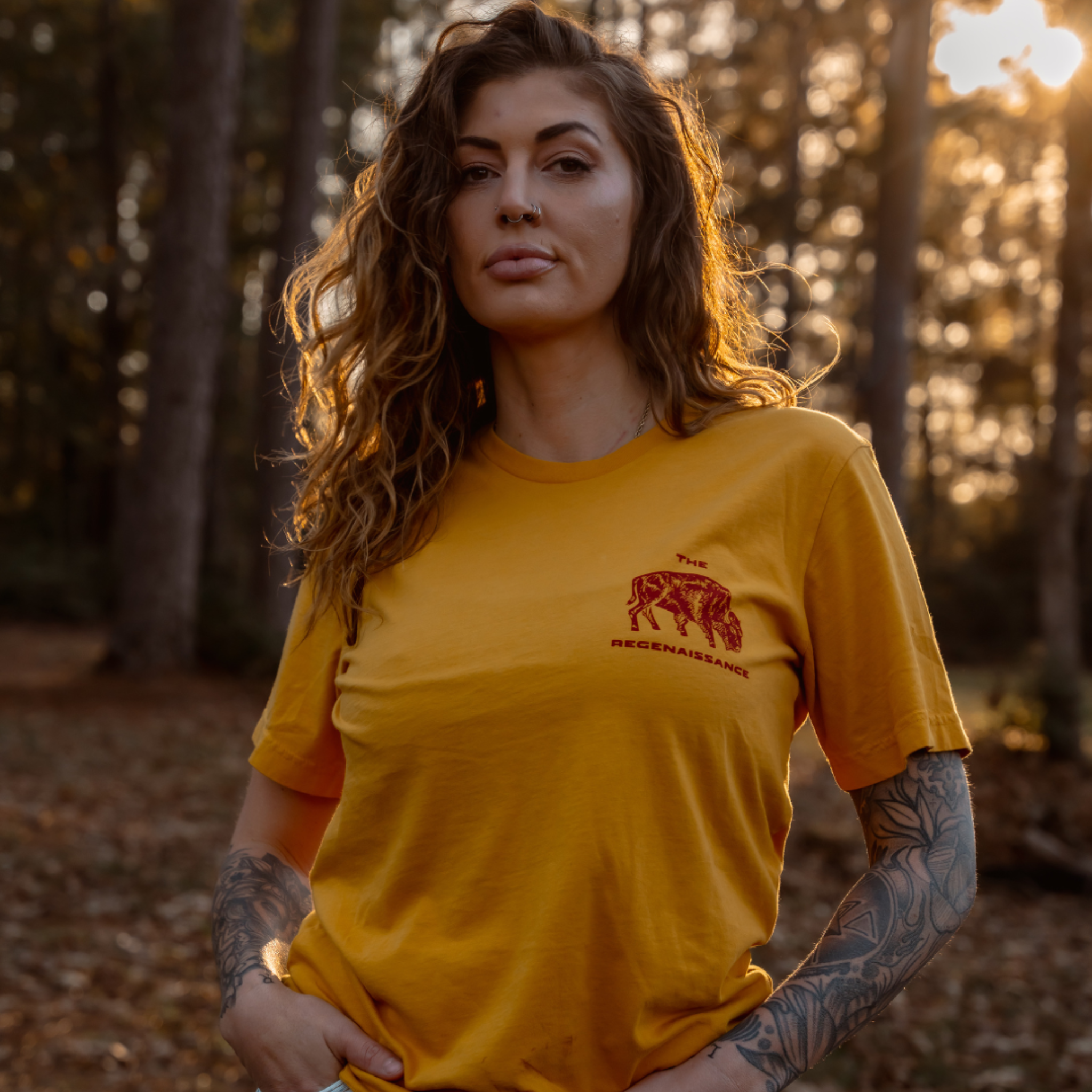 A woman with wavy hair and tattoos poses outdoors in a sunlit forest wearing the Farm Fresh Clothing Bison Tee, a mustard organic cotton T-shirt featuring red The Renaissance text and an illustration of two lions.