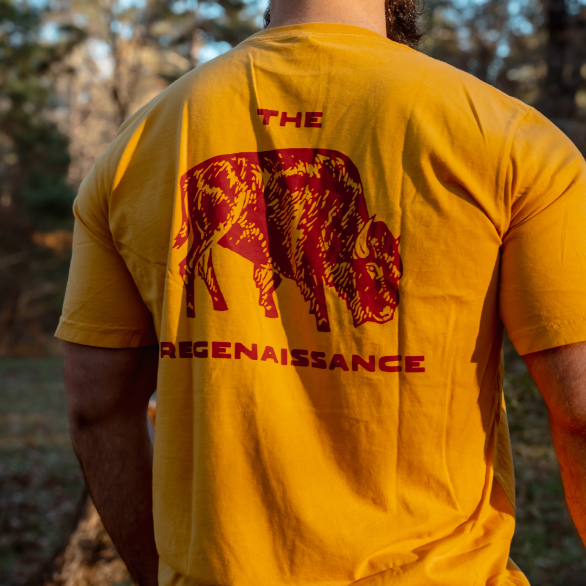 Outdoors, someone wears the Farm Fresh Clothing Bison Tee, a yellow organic cotton shirt with a red bison graphic and THE REGENAISSANCE on the back. Sunlight streams through trees in the background.