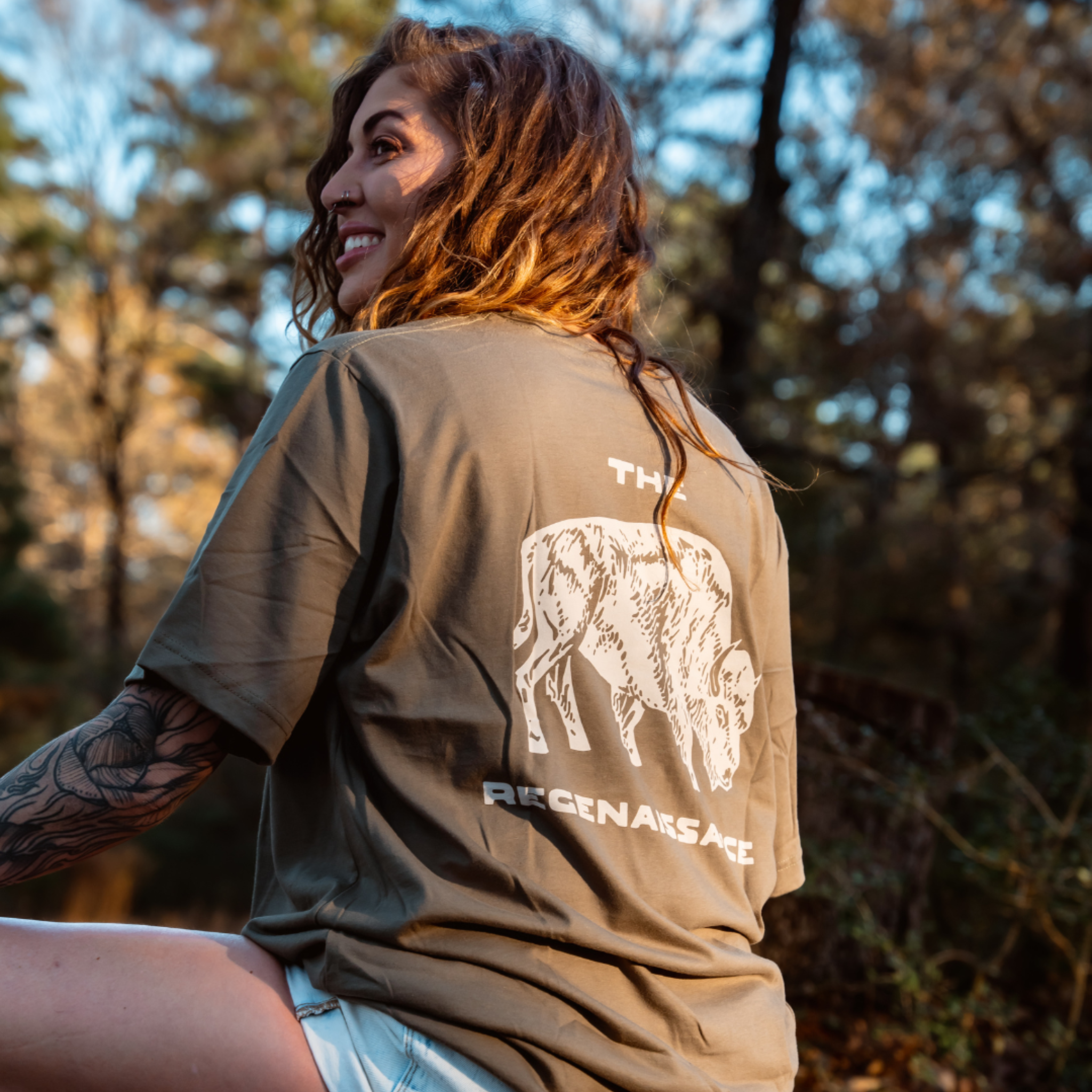 A woman with wavy hair and a tattooed arm sits outdoors, smiling in a Farm Fresh Clothing Bison Tee made from organic cotton. Sunlight filters through trees, reflecting the spirit of regenerative agriculture and THE REGENAISSANCE on the back.