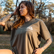 A woman with long hair and tattooed arms stands outdoors wearing Farm Fresh Clothing’s Bison Tee in olive green, made from organic cotton, as sunlight filters through the trees in the background.