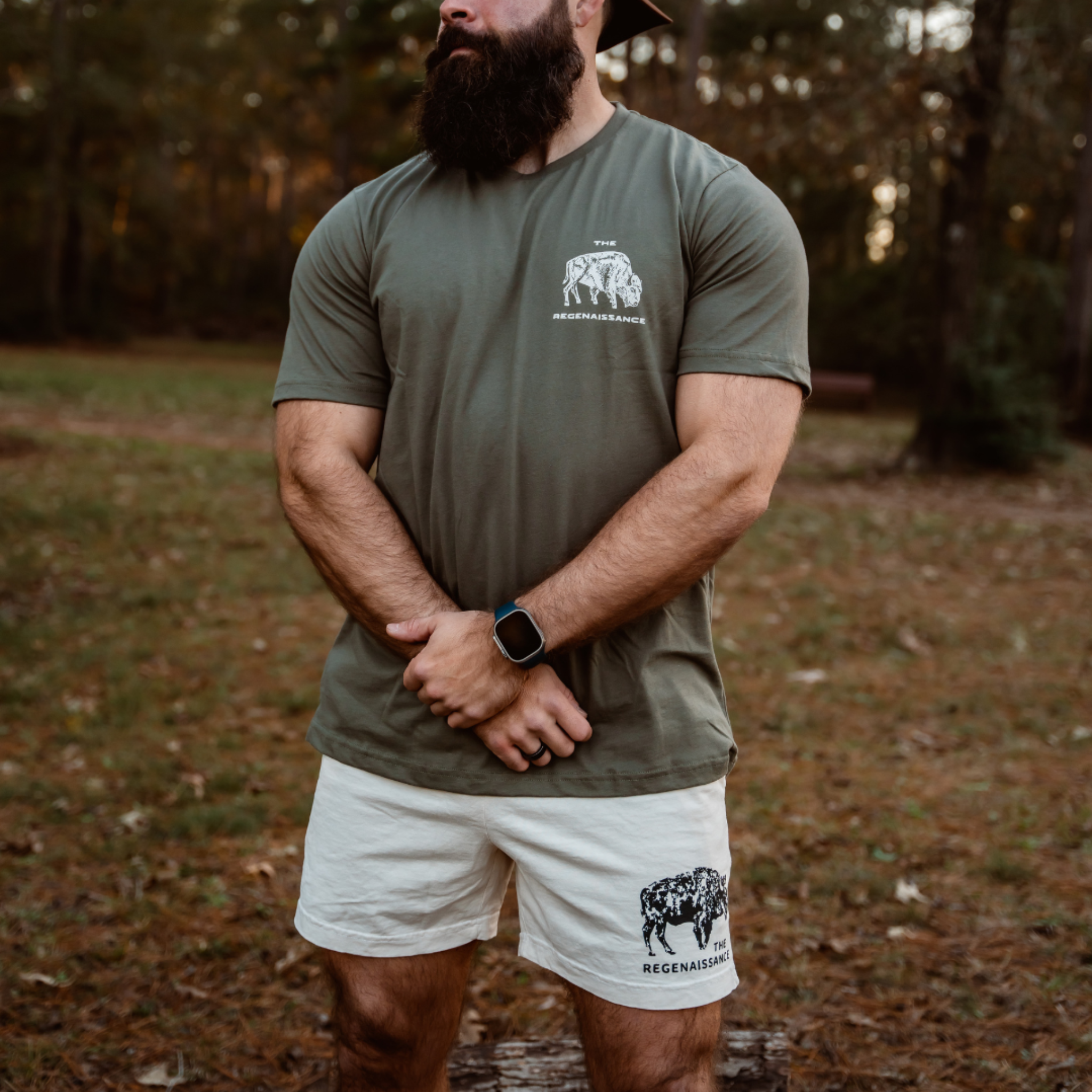 A bearded man stands on grass with crossed arms, wearing the Farm Fresh Clothing Bison Tee in Rosemary and white shorts featuring a black logo. He also wears a smartwatch. Trees and foliage appear in the background.