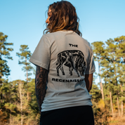 A tattooed individual with long hair stands outdoors in a Farm Fresh Clothing Bison Tee—an organic cotton shirt featuring a grazing bison and THE REGENAISSAN, framed by pine trees and blue sky, celebrating regenerative agriculture.
