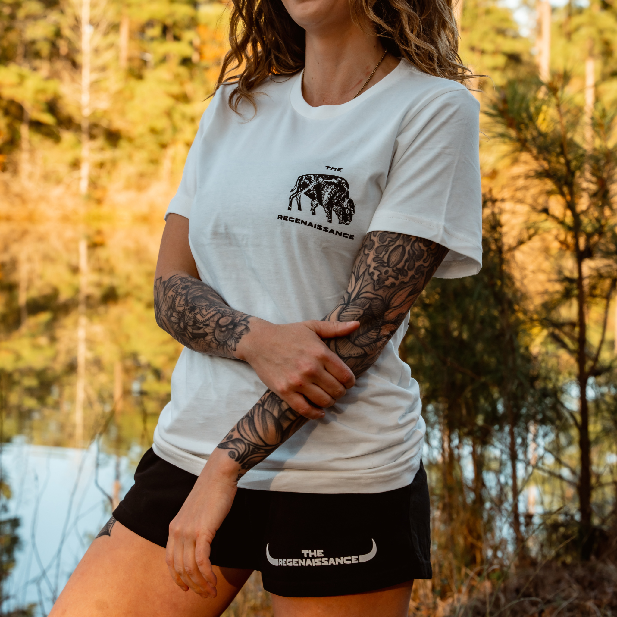 A person with long hair and tattoos on their left arm stands outdoors by a lake, wearing a white Farm Fresh Clothing Bison Tee and black shorts. Trees and water are visible in the background.