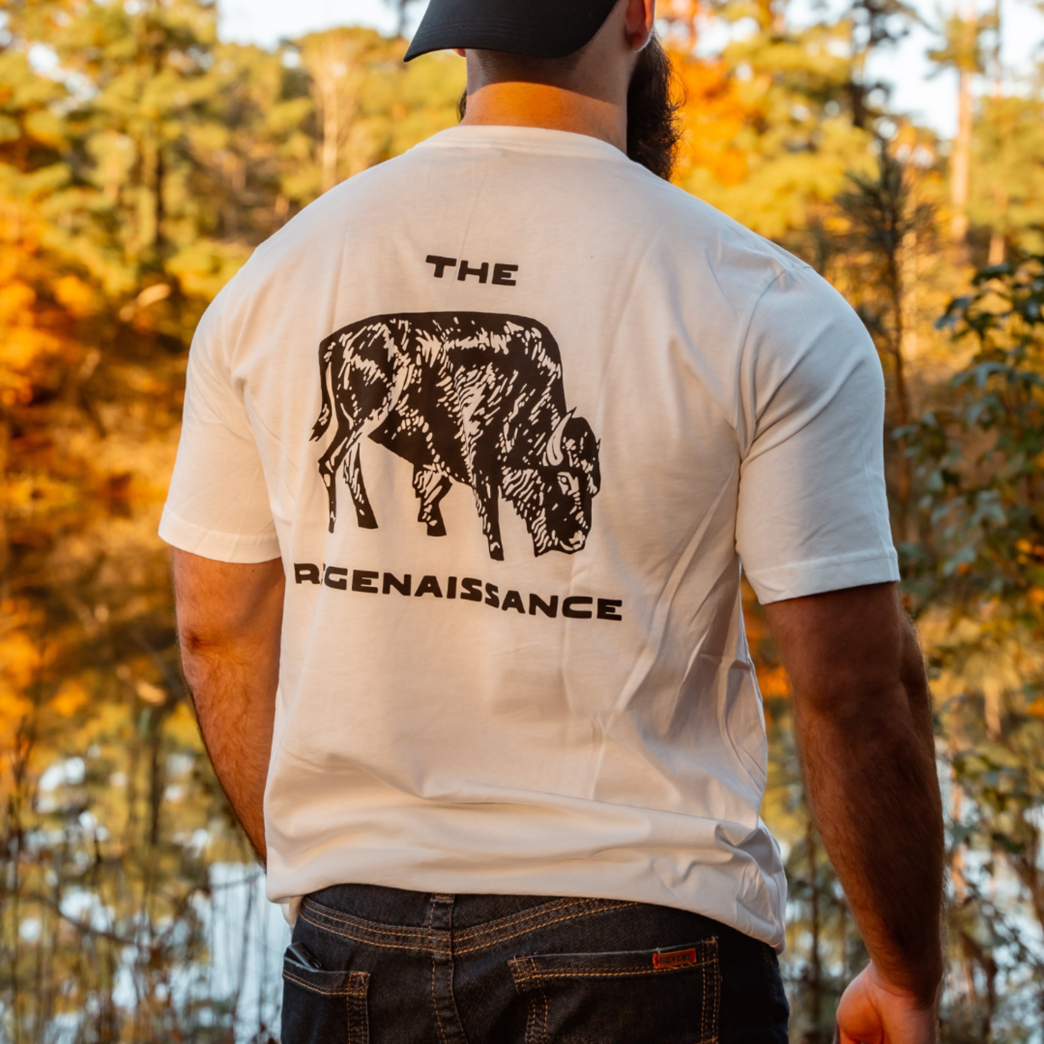 A man in the Farm Fresh Clothing Bison Tee, made from organic cotton with a black bison illustration and “THE ARGENAISSANCE,” stands outdoors by a lake amid autumn trees.