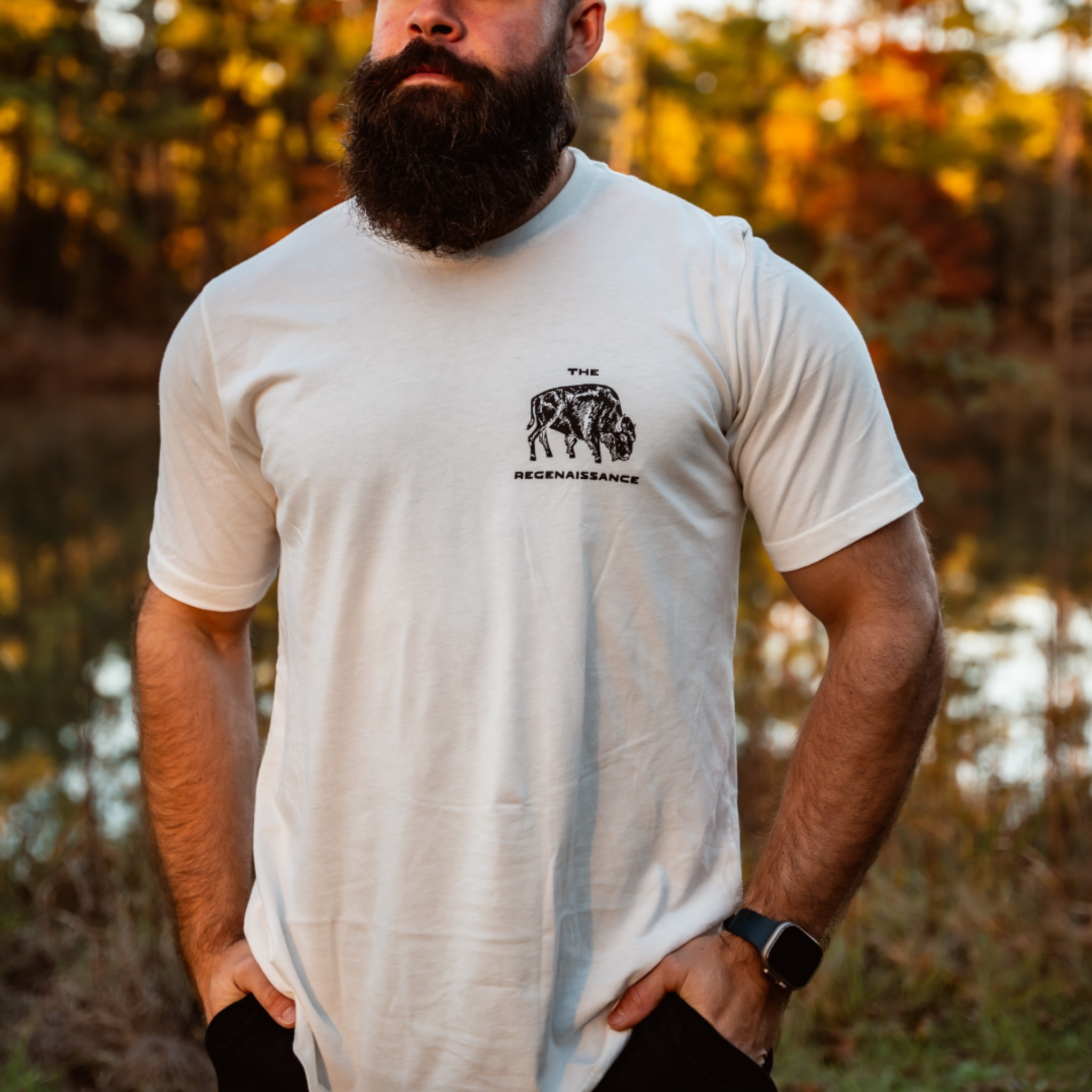 A bearded man stands outdoors by trees and a pond wearing an eggshell Bison Tee from Farm Fresh Clothing, featuring THE BECENAISSANCE text and a chest graphic.