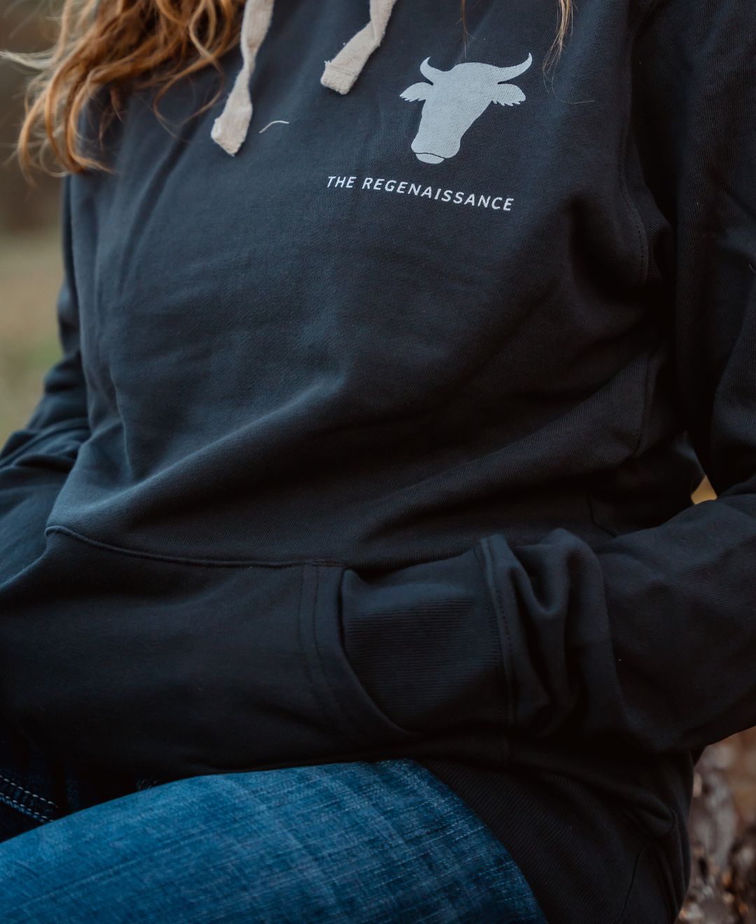 A person wears a Farm Fresh Clothing Raw is Law Hoodie with a bull head graphic and THE REGENAISSANCE text on the chest, paired with blue jeans. Hands in the pocket, they embody food freedom.