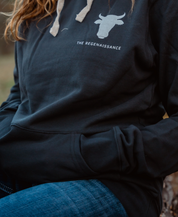 A person wears a Farm Fresh Clothing Raw is Law Hoodie with a bull head graphic and THE REGENAISSANCE text on the chest, paired with blue jeans. Hands in the pocket, they embody food freedom.