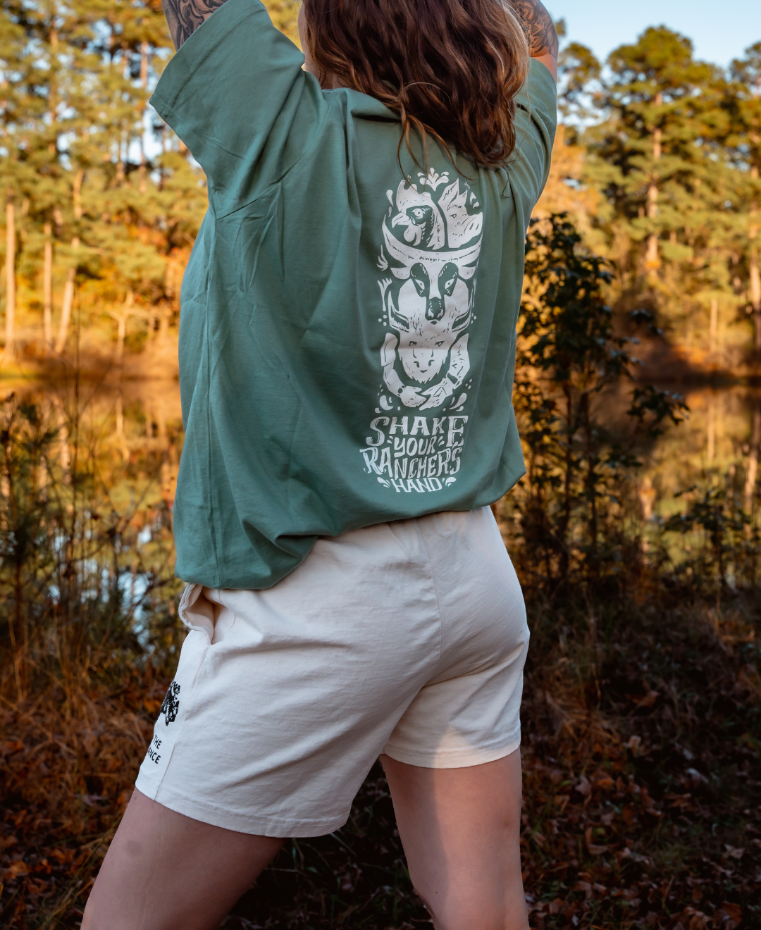 Outdoors, a person raises their arms wearing a green t-shirt with a skull-in-cowboy-hat graphic reading "SHAKE YOUR RANCHER HANDS," paired with Los Angeles Apparel Women's Bison Shorts. Trees and a pond reflecting regenerative agriculture are behind them.