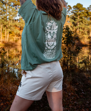 Outdoors, a person raises their arms wearing a green t-shirt with a skull-in-cowboy-hat graphic reading "SHAKE YOUR RANCHER HANDS," paired with Los Angeles Apparel Women's Bison Shorts. Trees and a pond reflecting regenerative agriculture are behind them.