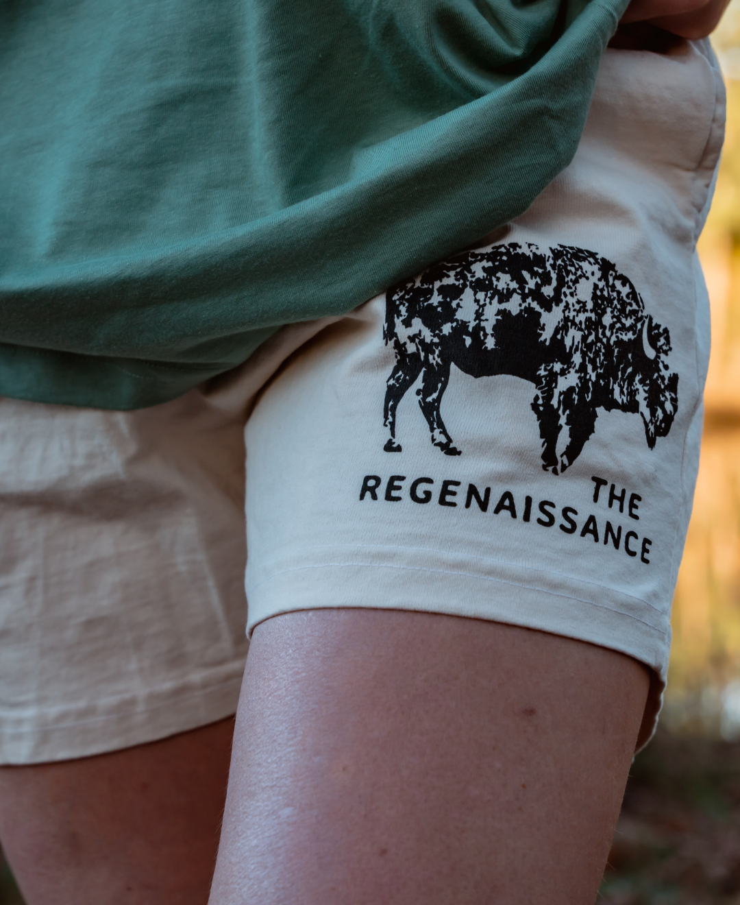 A person wears Los Angeles Apparel Women's Bison Shorts with a black bison graphic and THE REGENAISSANCE text, inspired by regenerative agriculture, partially covered by a green shirt.