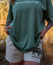 A person stands outdoors in a green The Regenaissance T-shirt and Los Angeles Apparel Women’s Bison Shorts, featuring a black graphic and text. They hold the shirt’s hem, with trees and sunlight in the blurred background evoking regenerative agriculture.