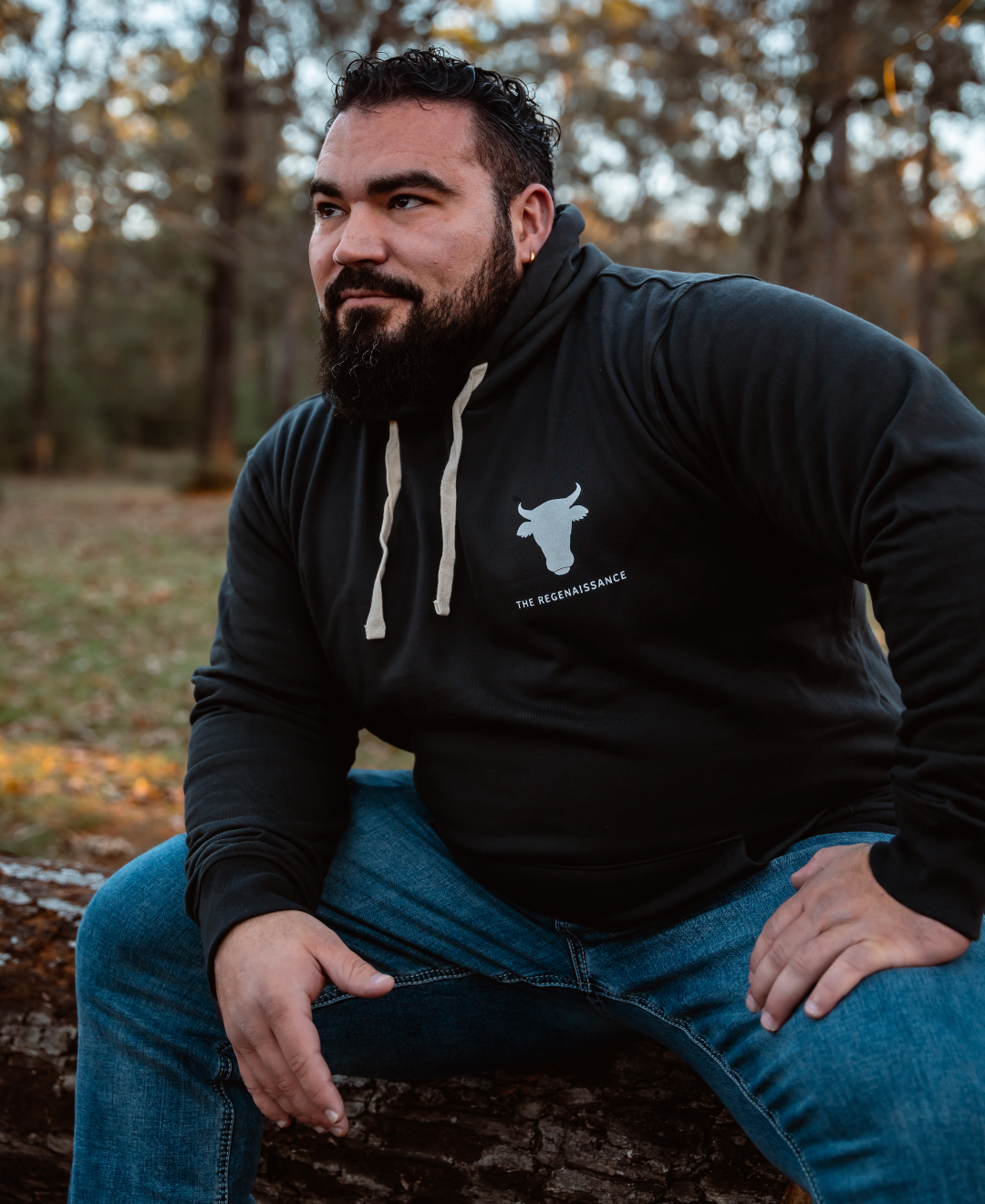 A bearded man in a gunpowder Raw is Law Hoodie from Farm Fresh Clothing and blue jeans sits on a fallen log outdoors, relaxed and enjoying nature’s peace and the spirit of food freedom as sunlight filters through the forest behind him.