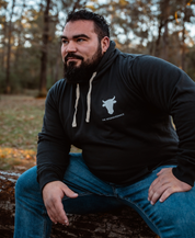 A bearded man in a gunpowder Raw is Law Hoodie from Farm Fresh Clothing and blue jeans sits on a fallen log outdoors, relaxed and enjoying nature’s peace and the spirit of food freedom as sunlight filters through the forest behind him.