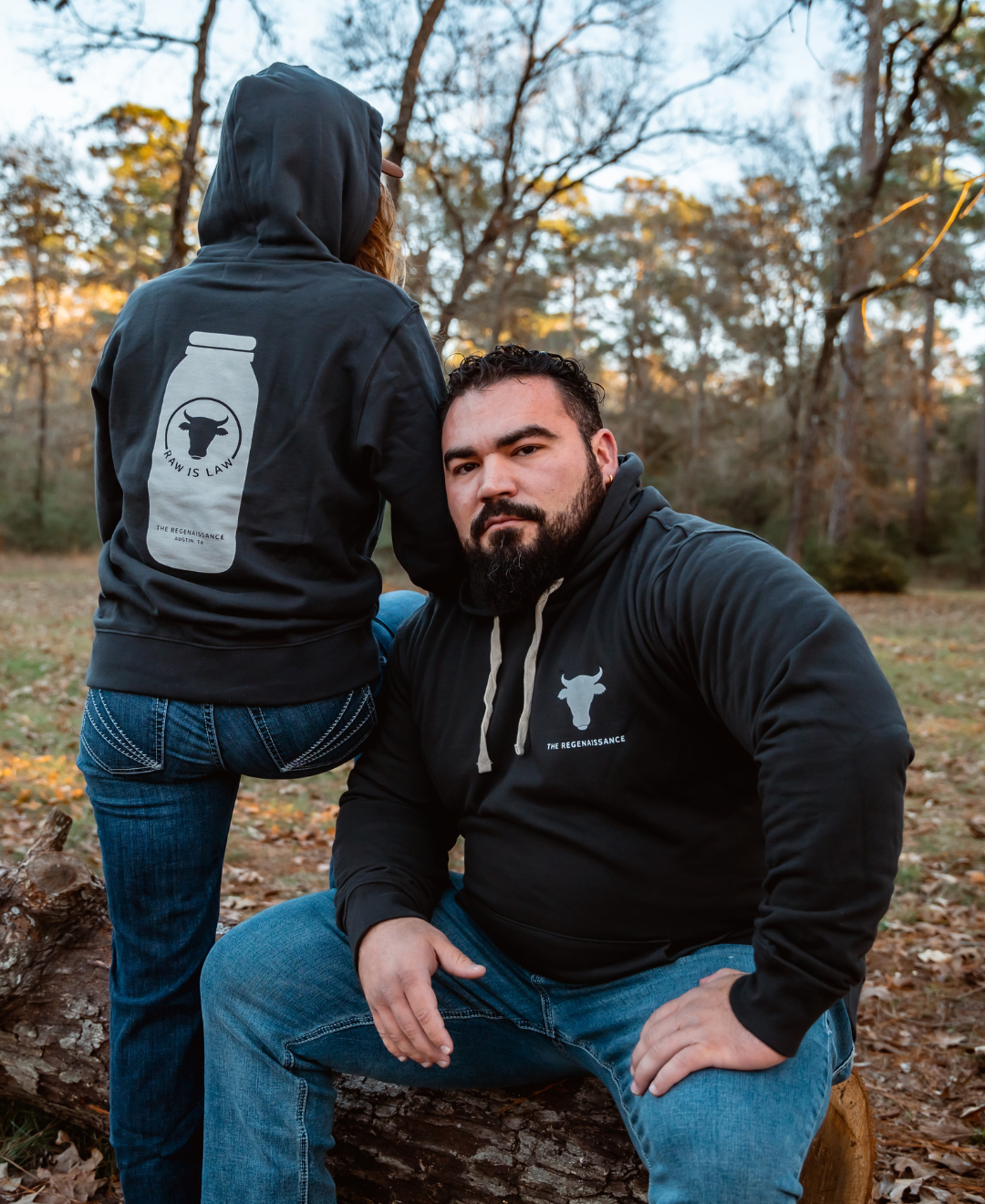 A man outdoors wears a black Farm Fresh Clothing hoodie with a white cow logo, ideal for food freedom and raw milk fans. Next to him, another person sports the Raw is Law Hoodie featuring a jar graphic and the phrase “How Is Lunch?”.